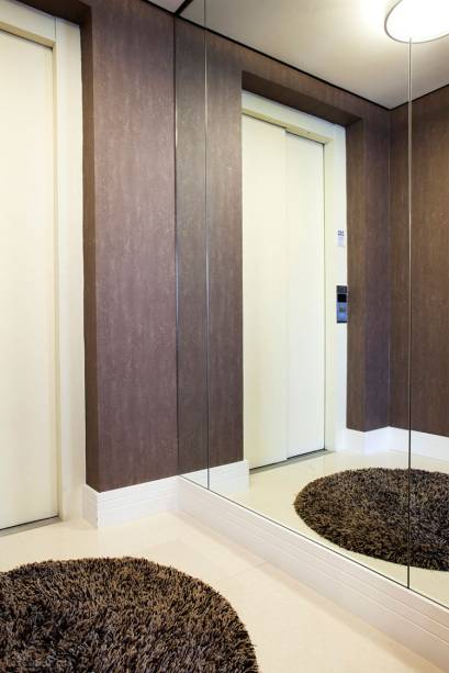 Hall de Entrada residencial com piso Mármore Crema Marfil,  MDF branco no rodapé e papel de parede marrom. Tapele de nylon e luminária embutida redonda em metal. Projeto de Lucia Helena Bellini.