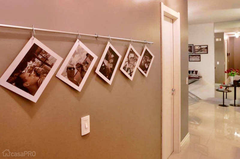 Com uma ideia super simples, porém original, este corredor acabou ganhando uma graça toda especial. Fotos impressas em adesivo vinílico texturizado foram aplicadas em plaquinhas de poliestireno presas por ganchos em um varão cromado fininho, daqueles que utilizamos em cortinas delicadas. Projeto de Aline Dal´Pizzol.