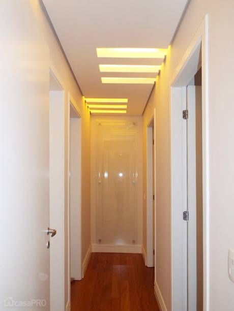 A circulação íntima deste apartamento de 123 m² teve como diferencial a iluminação e o elemento decorativo na parede da profundidade. Com 3,55 m de comprimento e 0,90 m de largura, o corredor possui as portas de acesso ao banho social, escritório, quarto de hóspedes e suíte do casal. O ponto de destaque principal é a iluminação decorativa efetuada com rasgos no forro de gesso pintado com tinta látex fosca. O outro ponto de destaque está no final do corredor: a única parede livre do ambiente ganhou um painel de vidro translúcido fixado com espaçadores cromados. Projeto de Vanessa Cassola Arquitetura.