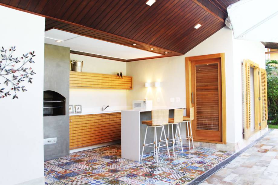 Paredes em textura cor neutra, churrasqueira em cimento queimado e armários em madeira. Nada poderia competir com o alegre colorido do ladrilho hidráulico utilizado no piso deste Espaço Gourmet. A bancada em quartzo branco também colaborou dando a leveza necessária para o piso ser o destaque principal deste ambiente. Os pontos em led do piso, assim como a iluminação das arandelas e fita em led abaixo do armário, deram um ar intimista para a programação noturna. Projeto de Paula Müller.