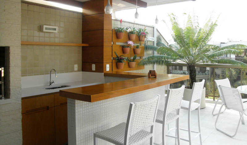 A proprietária desta cobertura solicitou à designer Ana Lucia Nunes Gomes um ambiente com churrasqueira, mas que não fosse rústico. A designer então optou por fazer uma bancada de Silestone branco e a churrasqueira de material cimentício da Solarium. Um pequeno jardim vertical abriga vasinhos de flores e aproveita a luz natural.