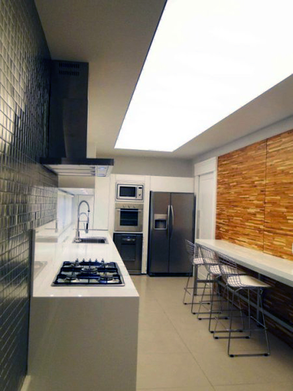 Misturar texturas também pode acontecer em cozinhas em estilo corredor. Este projeto de Michelle Donida utiliza pastilhas metálicas de um lado e madeira do outro. Para não sobrecarregar o ambiente, a marcenaria foi feita toda em branco.