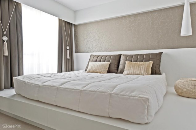 A cama em estilo tablado ocupa toda a extensão deste quarto assinado por Renata Mueller. A marcenaria branca vai até metade da parede, que foi complementada com um papel texturizado. Repare que o tecido do travesseiro se repete na cortina.