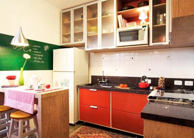 A cozinha projetada pela arquiteta Thais Lenzi Bressiani utiliza o vermelho no armário e em diversos acessórios . Na parede, uma tinta que imita lousa é utilizada para anotar recados e receitas. Para não pesar no ambiente, os armários ganharam portas translúcidas.