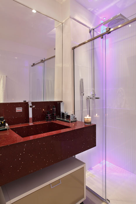 Para o morador solteiro, a designer Ana Lucia Martins projetou um banheiro clean, com pequenos elementos em destaque. Por isso, a escolha da bancada de eco quartzo na cor vinho e iluminação do boxe em LED azul.
