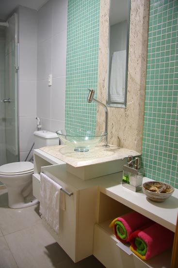O banheiro de 3,6 m2, criado por Larissa Vinagre, tem detalhes de pastilhas de vidro verde água e uma bancada de mármore travertino. As paredes são revestidas com cerâmica branca e o piso é porcelanato esmaltado. Como o banheiro faz parte da área social do apartamento, a arquiteta optou por dar a ele uma cara de lavabo, embora possa ser usado para banho.