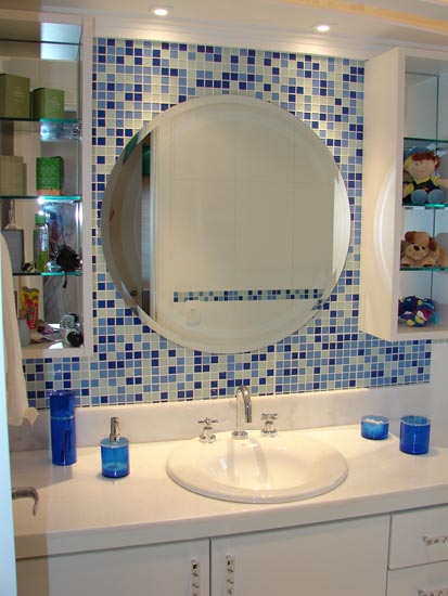 As pastilhas de vidro em azul e branco combinadas com o espelho redondo e com os nichos laterais brancos  que tem prateleiras de vidro  dão o charme deste banheiro assinado por Lívia Gobetti. A bancada tem um tampo de mármore branco. A iluminação é feita através de lâmpadas dicróica.