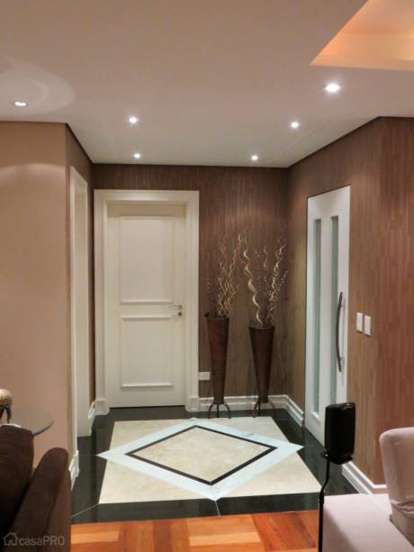 Hall de entrada com piso de granito e mármore, paredes revestidas com papel de parede, porta laqueada em branco com detalhes em vidro e cachepôs com vegetação seca decorando o ambiente. Projeto de Daarna Arquitetura & Interiores.