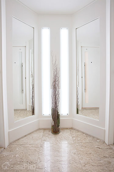 Hall social residencial. Paredes revestidas em MDF branco liso, com iluminação em led embutido, piso mármore travertino. Projeto Rose Ferraresi.
