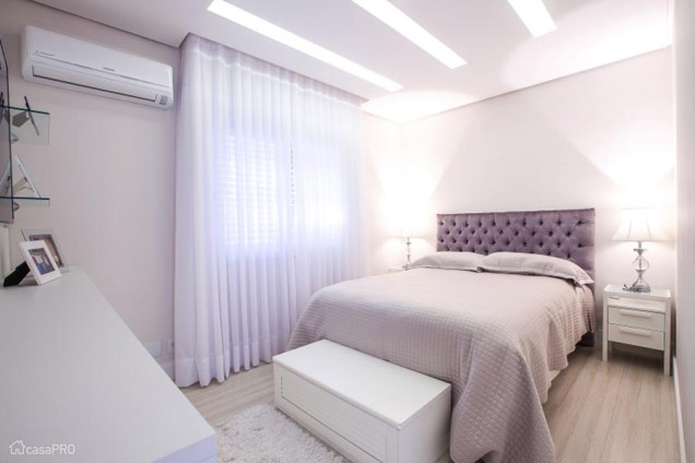 As paredes claras dão uma sensação de amplitude para este quarto projetado por Camila Chalon. A cabeceira é de capitonê roxo, o que dá um tom romântico ao ambiente.