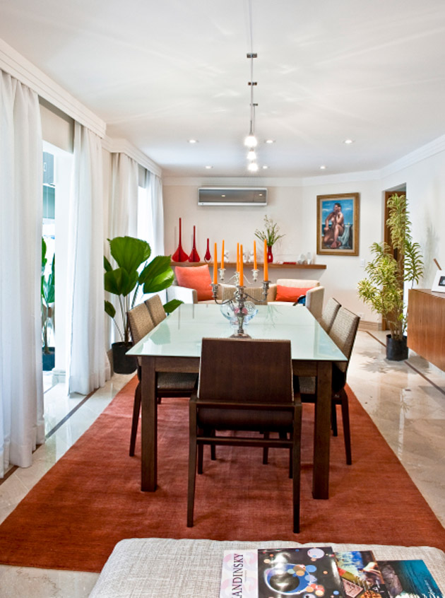 A mesa retangular tira proveito da sala com planta estreita. Dois tipos de iluminação destacam os móveis. Projeto de Rosana Pedroso.