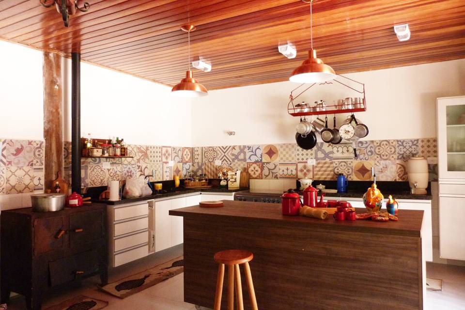 A cozinha de 53 m&sup2; &eacute; o destaque desta casa projetada por Elizabeth Berlato. O ambiente possui fog&atilde;o a lenha, com uma bancada no centro para ajudar no preparo dos alimentos. As paredes trazem acabamento em porcelanato que imitam ladrilho hidr&aacute;ulico.