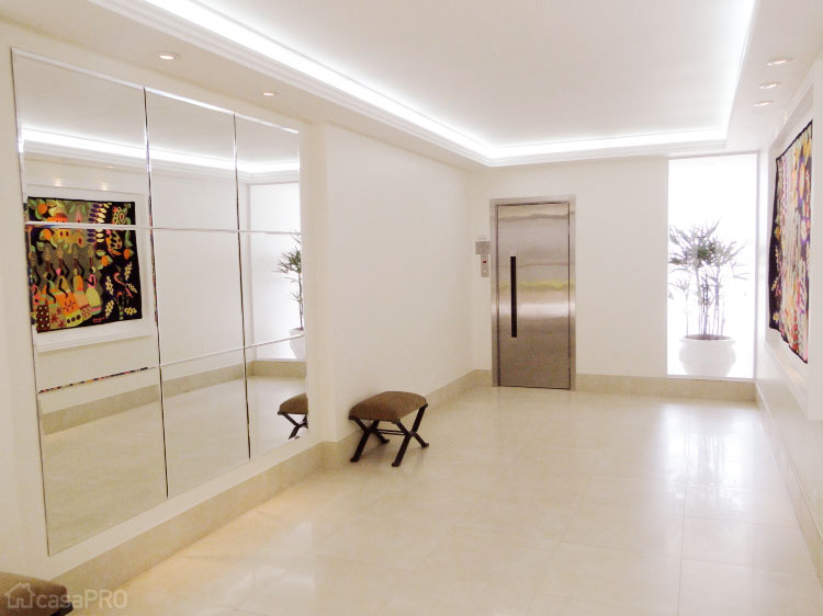 Hall social com piso porcelanato 60 x 60 cm, parede em drywall revestida com painel de espelho, iluminação com fitas de led embutidas na sanca de gesso e iluminação direta na tapeçaria. Projeto Rose Ferraresi.