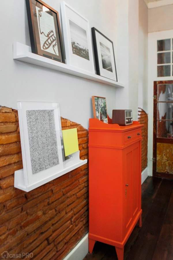No corredor, sobre prateleiras apoiam-se quadros com fotos e ilustrações de artistas catarinenses que compõem com a cômoda laranja também reformada. Parede com tijolo aparente. Projeto do Studio UM Interiores.