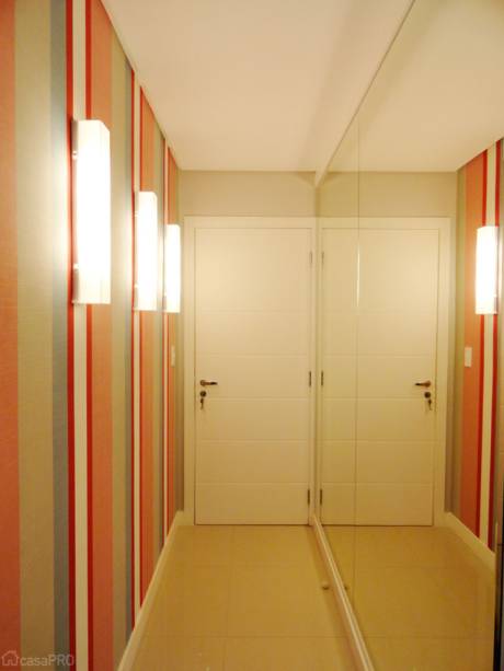 Hall de apartamento pensado para receber com aconchego e sofisticação, por isso o uso do tecido e das arandelas. O painel de espelhos amplia visualmente o espaço e diminui a sensação de corredor. As paredes receberam tecido para decoração Karsten, tinta Reco-Reco Suvinil e painel com espelho Cristal. O piso foi revestido com porcelanato polido 60 x 60 cm da Portinari. Iluminação: arandelas em acrílico feitas sob medida pela MZ Lustres. Projeto do Frente & Verso Arquitetura.