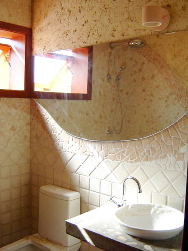 Para o banheiro da sala de jogos, a arquiteta Carla Prado Dadazio escolheu criar um ambiente mais rústico. A parte superior da parede recebeu um reboco grosso e um gel de textura, conversando com a bancada de concreto com pedras de seixo salpicadas pela superfície.