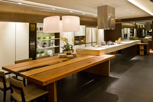 CASACOR Brasília - No centro da cozinha, projetada pela arquiteta Walléria Teixeira, a bancada feita de Corian material feito a partir de resina abriga o cooktop e a torneira gourmet. A coifa de design retangular deixa a cozinha mais sofisticada. Uma mesa de madeira dá espaço para refeições e convivência. A cozinha ampla de 70 m² possui armários com portas brancas e espaços para estantes.