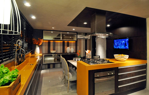 CASACOR Pernambuco - Assinada pelo arquiteto André Dantes, a Cozinha do 2º pavimento da CASACOR Pernambuco inova com os balcões de silestone em laranja. Na parede ao fundo, um painel de madeira de diversas tonalidades foi montado. A cozinha é bem equipada com eletrodomésticos Falmec, Exclusive line e Latina. O teto na cor de concreto é brilhante e resistente a umidade.