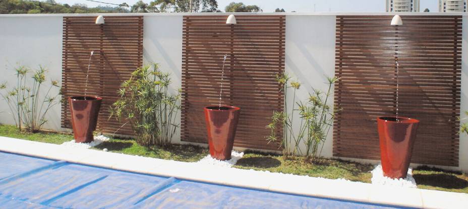 Com apenas 50 cm de terra entre o piso da borda da piscina e o muro, a designer de interiores Fabiana Terra criou um projeto de paisagismo que inclui três fontes: uma bombinha de aquário jorra água em vasos vietnamitas vermelhos, dispostos em frente a um painel de madeira cumarú envernizado e sobre pedriscos brancos.  Projeto localizado em um condomínio fechado na cidade de Santana do Parnaíba (SP).