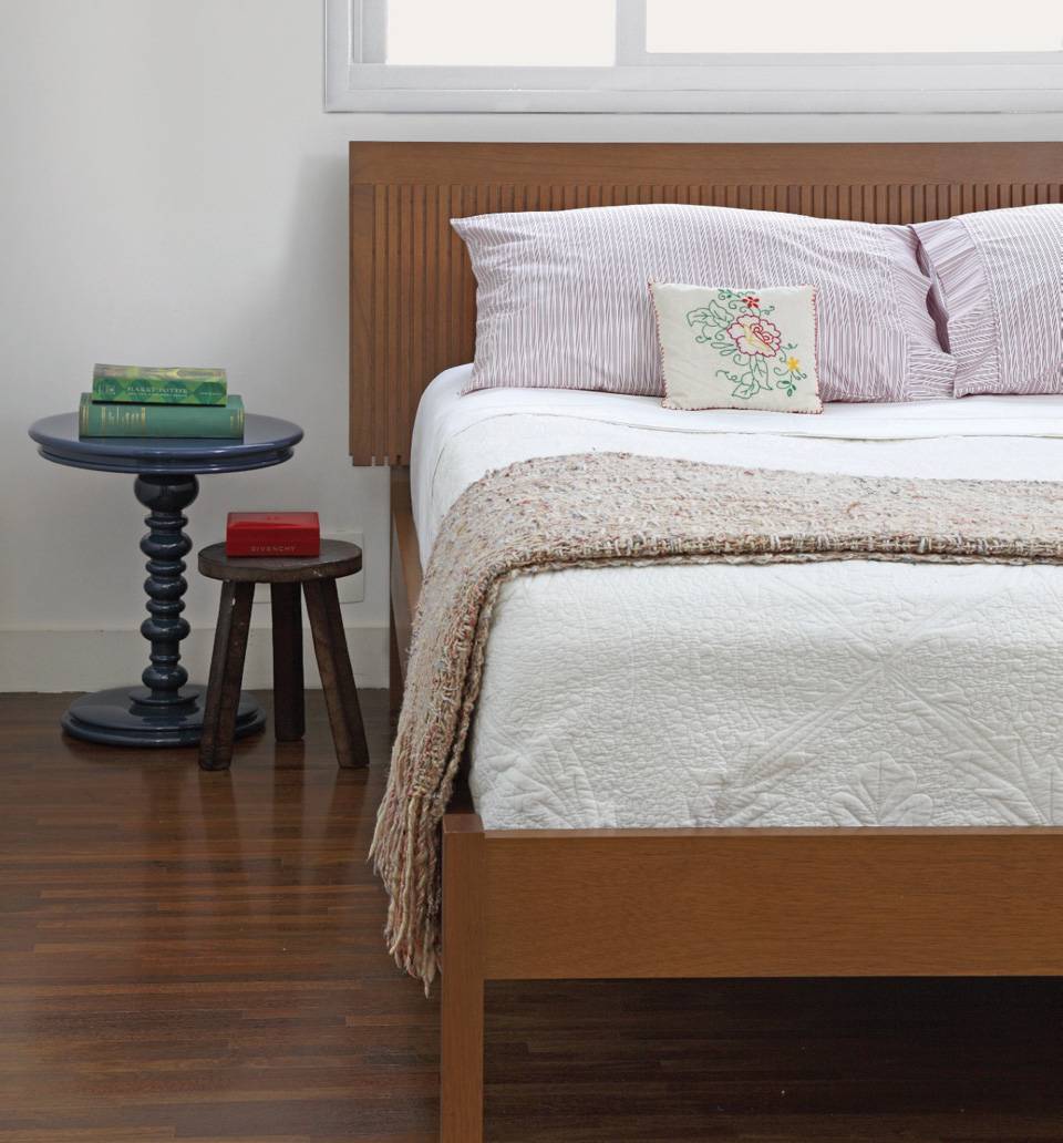 O fundo branco e a cama com estrutura e cabeceira de madeira permitem ousar em detalhes, como a mesa com tampo redondo pintada de azul que faz o papel de criado-mudo. O quarto &eacute; da arquiteta Carolina Zupiroll.