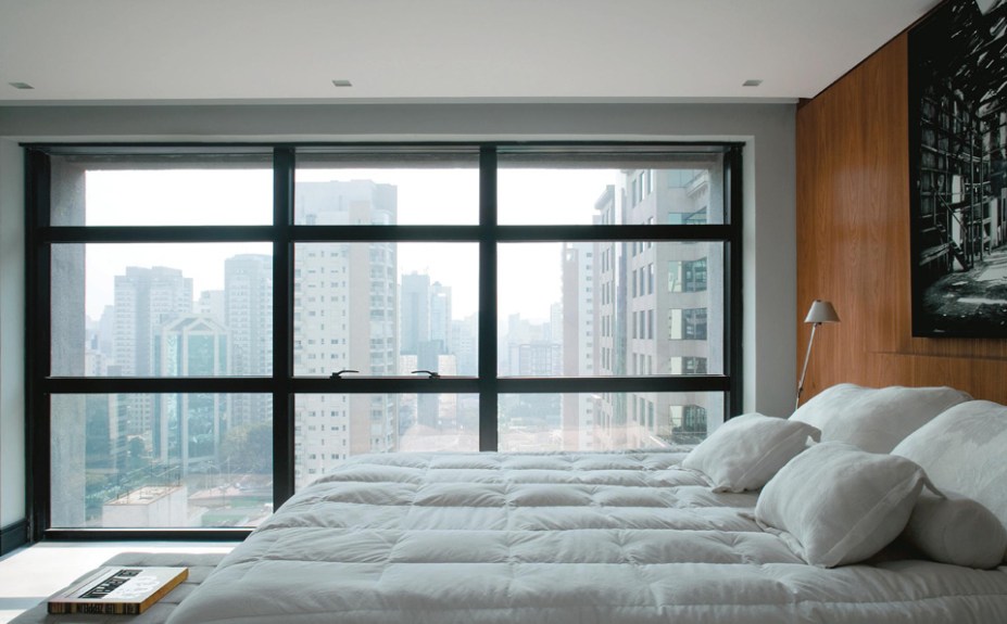 O skyline da capital paulista impera através da janela do quarto em preto e branco. Para deixar o espaço mais acolhedor, painéis de madeira (Marupá Móveis) revestem as paredes.