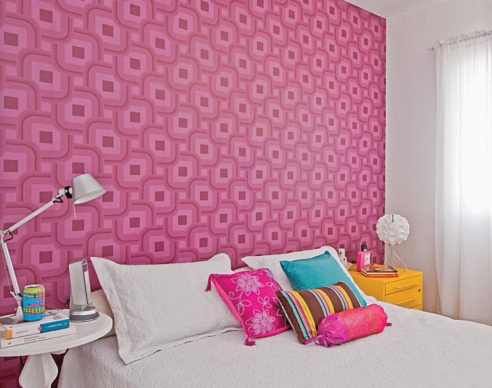 O papel de parede (cole&ccedil;&atilde;o Grafismo, linha Azulejo, da Bobinex) foi a base para escolher os demais elementos coloridos no quarto, projetado pela arquiteta Juliana Savelli. O rosa na parede e nas almofadas, e o amarelo da mesa lateral (Modelo Rolling, de a&ccedil;o. Tok & Stok) trazem alegria ao branco.