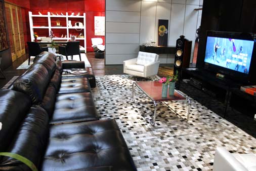 Além do grande sofá de couro preto, o tapete desta sala é todo feito de re...