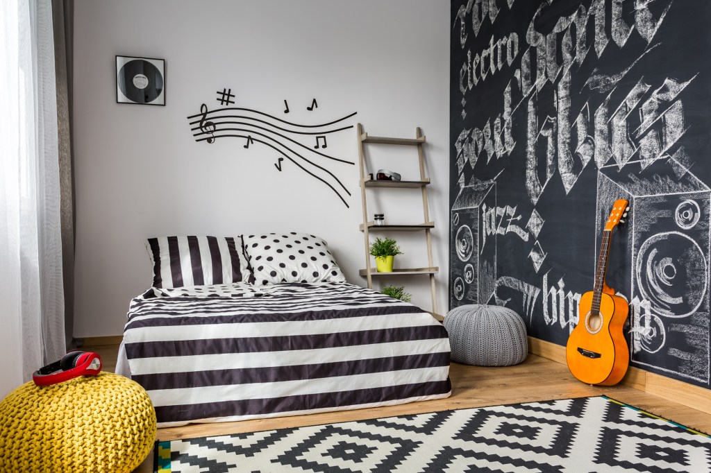 Quarto com decoração preta e branca, com cama listrada com um violão ao lado, tapete estampado. Notas musicais e um disco de vinil na parede atrás da cama. Parede de lousa com ritmos musicais escritos em giz.
