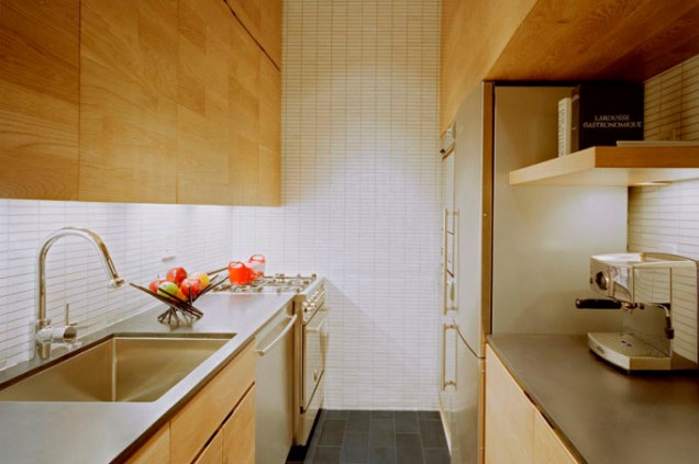 A madeira clara e azulejos bem pequenos, brancos, compõem esta cozinha em formato de corredor. A madeira conecta o espaço aos cômodos ao lado, de design similar. Ela também abriga os eletrodomésticos de aço inoxidável em armários que vão até o teto.