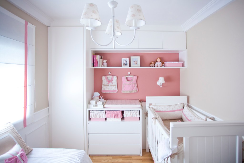 Quarto pequeno com berço e cama brancos. Na parede, da lado esquerdo, um armário até o teto, ao lado uma cômoda com trocado a frente de uma parede rosa, com uma prateleira e armario com quatro portas próximo ao teto