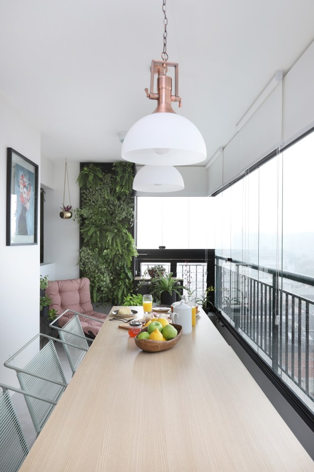 A varanda, como continuidade da sala, recebeu uma mesa grande de marcenaria e serralheria e possui um cantinho relaxante, com poltrona e um jardim vertical.