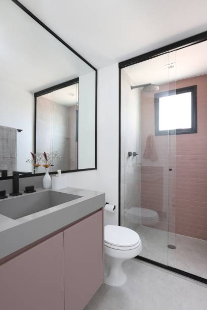 O banheiro da suíte carrega nos revestimentos o piso com padronagem de cimento queimado. O rosa matte do lavabo é replicado na bancada e na parede do box, cujas moldura, esquadria, moldura do espelho e metais são na cor preta, contrastando com os tons leves e marcando suas formas.