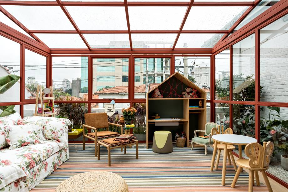 Varanda - Marina Linhares. Um gazebo de 39m², construído a partir de uma estrutura metálica e em vidro, explorando a luz natural e integrado ao paisagismo de Gabi Pileggi, do Jardineiro Fiel. Pensado para reunir a família em um momento de contemplação do horizonte, o espaço é pontuado por flores e plantas. O interior apresenta móveis confortáveis estofados com tecidos naturais e objetos de design. Uma estante com dois metros e formato de casinha abriga uma coleção de brinquedos.