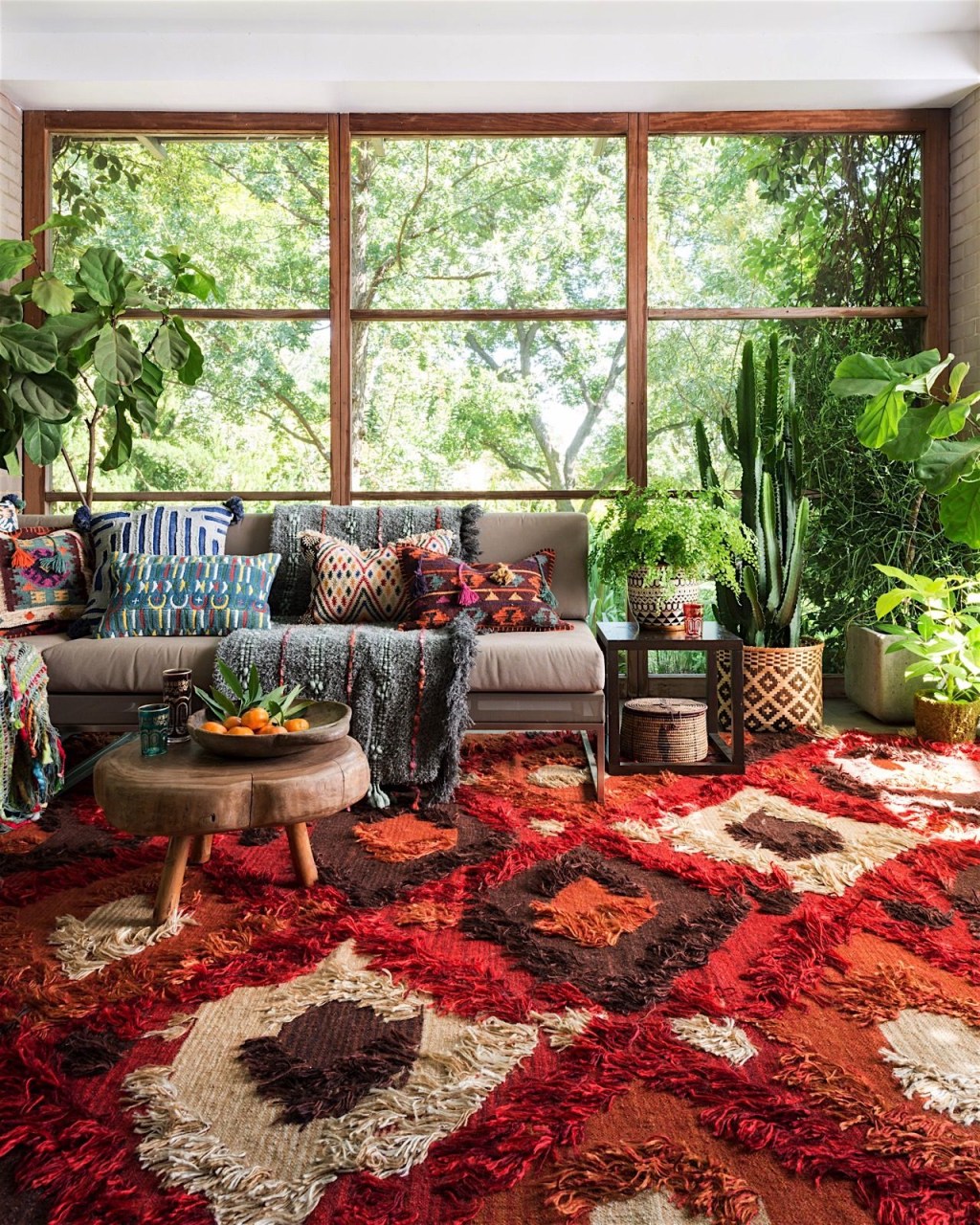 Decoraçnao estilo boho com tapete vermelho estamapado, sofá cinza com almofadas estampadas coloridas e muitas plantas