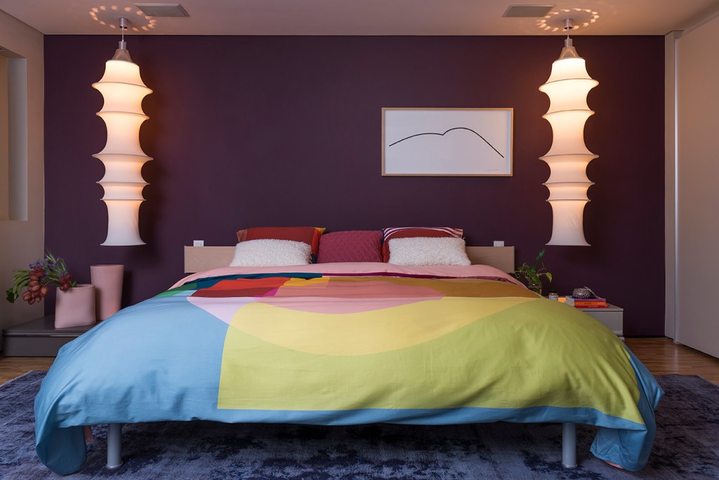 Quarto com parede roxa; roupa de cama; edredom colorido; luminárias pendentes; quadro; travesseiros coloridos