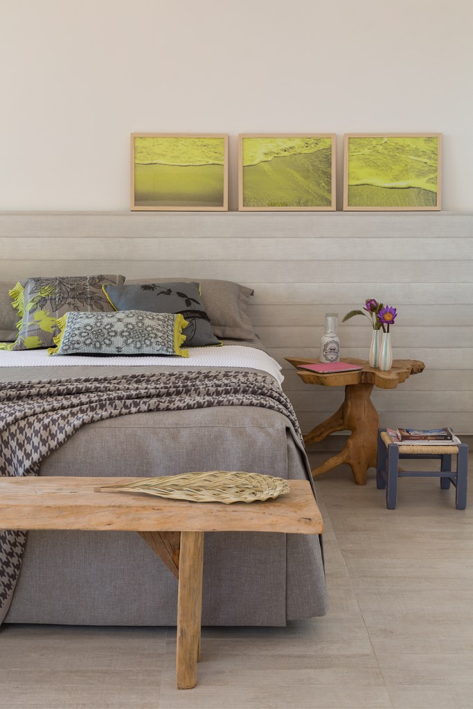 Quarto com meia parede; quadros; cama com roupa de cama cinza; mesinha lateral rústica de tronco
