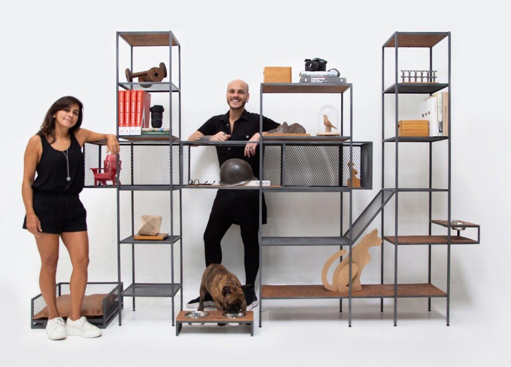 Acessórios de animais de estimação em estilo industrial criados pelos designers Pedro Galaso e Luciana Duque, que aparecem na imagem apoiados em uma estante modulada com objetos decorativos.