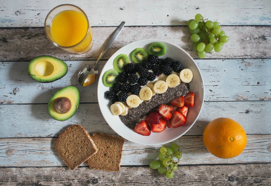 Pote com frutas picadas. Ao lado, há um copo de suco e duas fatias de pão integral.