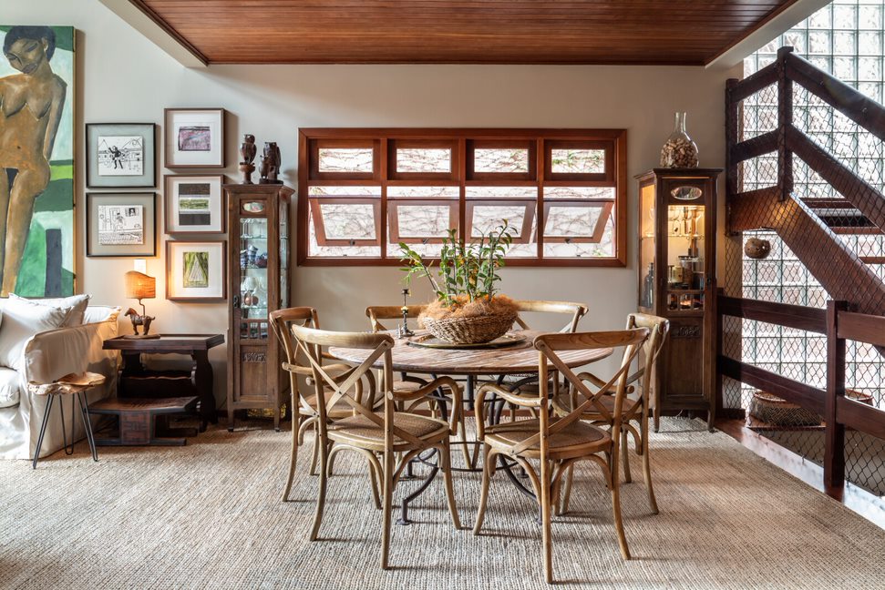 Sala de jantar com tapete de linho. Teto em madeira. Janelas em madeira ao fundo. No centro, mesa de jantar com cadeiras de madeira.