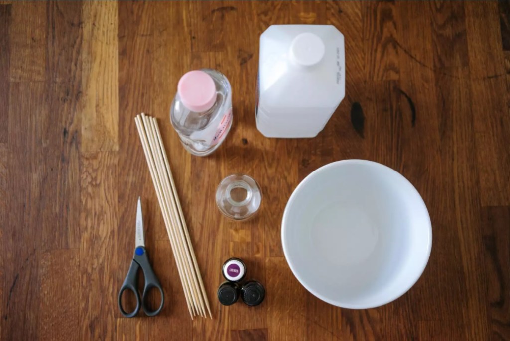 diy-difusor-aromaterapia-01-the=spruce-jorge-gamboa Mesa de madeira com produtos para fazer aromatizador. Tesoura, varetas, alcool, tigela, frasco de vidro e óleos. Todos vistos de cima.