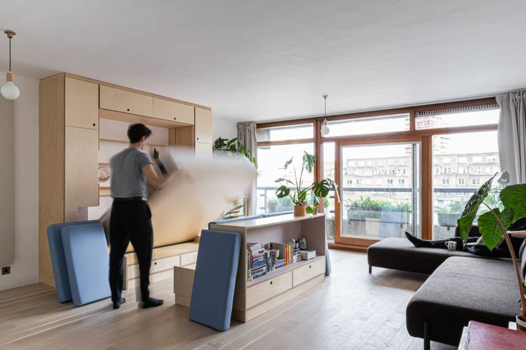 Flat com piso em madeira e grande janela. Homem abaixa colchão de móvel de madeira clara na parede.