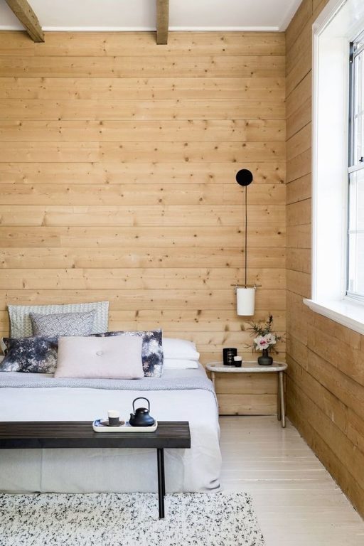 Quarto com parede de madeira e cama de casal branca com quatro almofadas. Luminária pendente pequena do lado direito funcionando como abajour