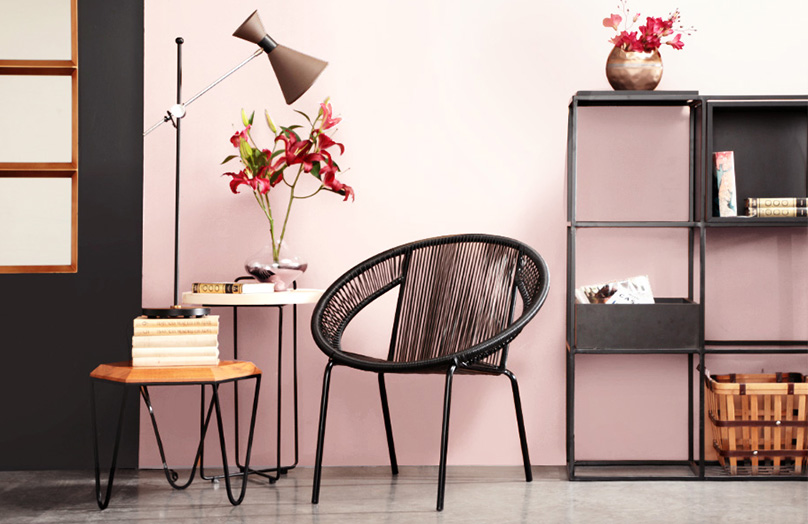 Ambiente com cadeira trançada preta, duas mesas de apoio, uma com flores rosa e a outra com um abajour. Estante preta ao fundo. Parede rosa clara