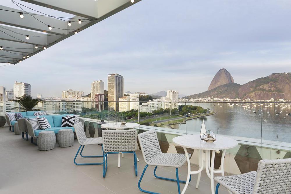 Cadeiras e mesas brancas no Rooftop do Yoo2, ao Fundo, a cidade do Rio De Janeiro, com o Pão de Açúcar e o mar.