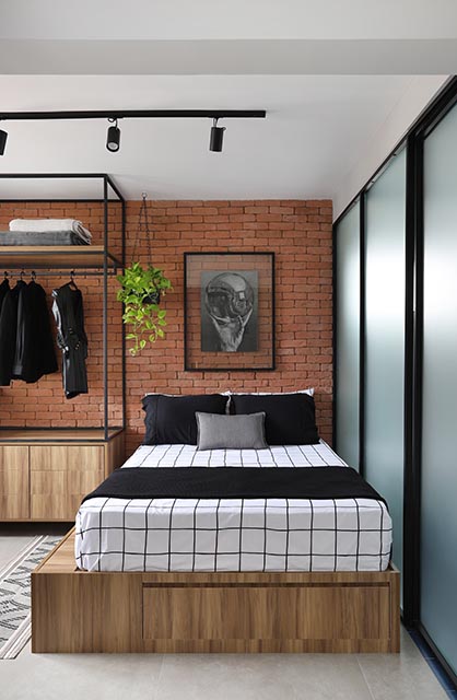Cama com lençol branco com linhas pretas, sobre uma base de madeira, no quarto com parede de tijolos laranja