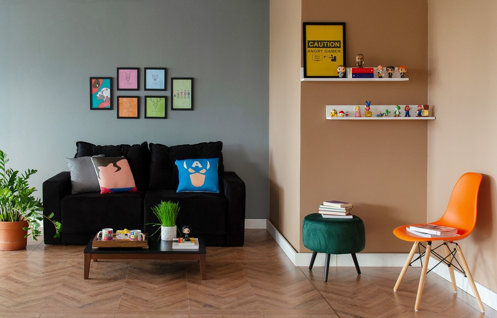 Sala com piso em madeira e sofá preto com almofadas de super heroi. Quadros na parede. Prateleiras com bonecos