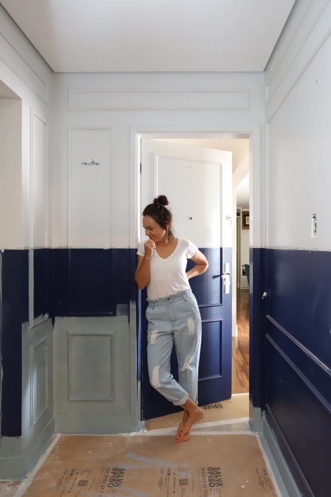 Mulher encostada no batente de uma porta que está semi aberta atrás delas. Ao seu redor, as paredes têm a metade infeior pintada de azul escuro, e a parte superior na cor branca.