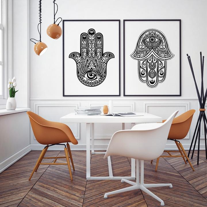 objetos-negatividade-mao-hamsa-06 Sala de jantar com piso em madeira e parede branca. Mesa com quatro cadeiras, duas laranjas e duas brancas. Ao fundo, dois quadros com mãos hamsa em preto e branco