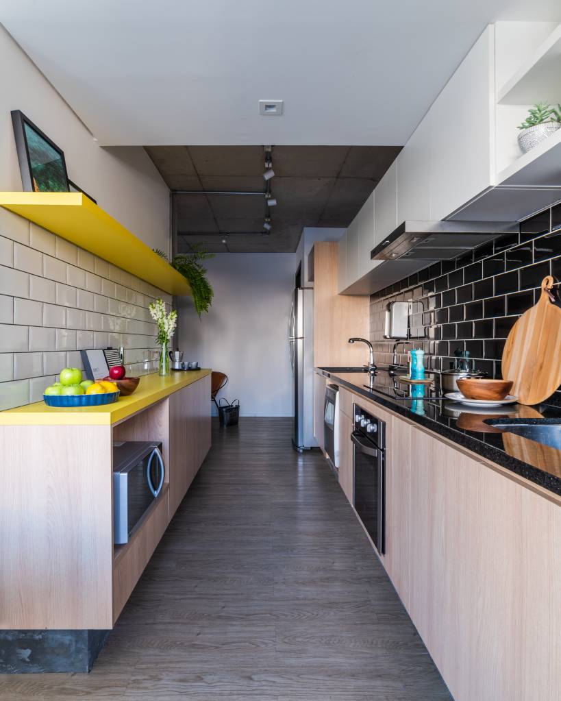 decor-industrial-marcam-feito-para-alugar-casa.com-5 cozinha indutrial com piso de madeira cinza, armários em madeira natural do dois lado no corredor, com o tampo amarelo do lado esquerdo e uma prateleira da mesma cor acima; do lado direito, os eletrodomésticos, com o tampo em pedra preta e armários aéreos branco acima. Nas paredes tijolinhos, do aldo esquerdo, brancos e do lado direito, pretos