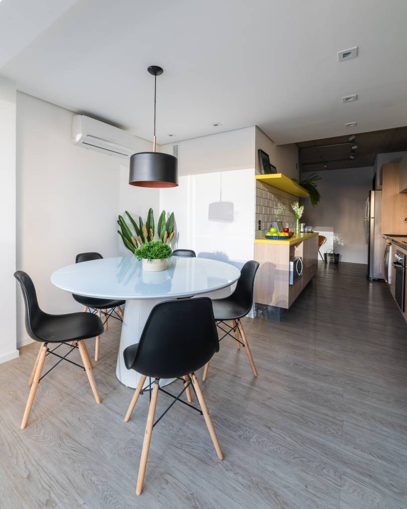 decor-industrial-marcam-feito-para-alugar-casa.com-6 Cozinha co estilo industrial, com mesa brnca e quatro cadeiras com assento preto e pés de madeira. O chão é de madeira cinza e as paredes são branca. Na mesa, um vaso pequeno com planta e acima dela um pendente com cúpula redonda vazada em cima e embaixo